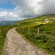 A dirt road cutting through green fields. The ocean is in the background with a rainbow faintly visible.